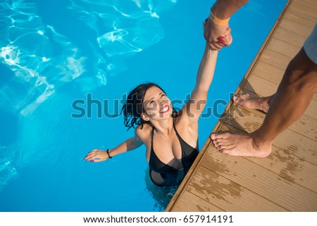 View from above of attractive girl in the swimming pool holding a man's hand trying to get out at resort. Sunny day