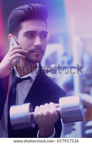 Handsome young man in a black suit, white shirt and tie talking on the phone with a dumbbell in hand in the gym