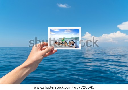 Hand holding a picture of happy big Asian family tourist on vacation with beautiful white clouds on blue sky over calm sea in background.