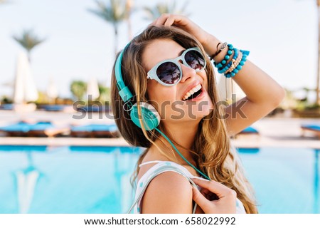 Close-up portrait of joyful laughing young lady in trendy bracelets posing with hand up near the open-air blue swimming pool. Cute girl in headphones enjoying favorite song on palm trees background
