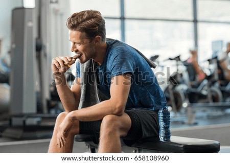 Sweaty young man eating energy bar at gym. Handsome mid guy enjoying chocolate after a heavy workout in fitness studio. Fit man biting a snack and resting on bench. Royalty-Free Stock Photo #658108960