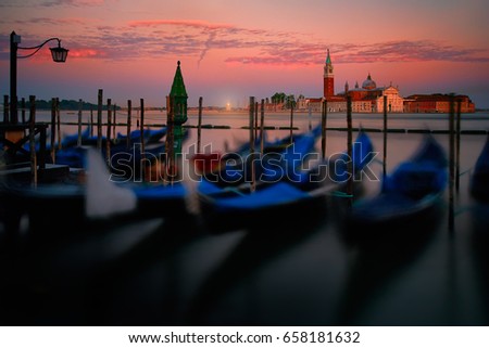 Artistic, long exposition photo of blurred gondolas moored by Saint Mark square with San Giorgio di Maggiore church and lighthouse in the background. Red colorful dusk. Tourist hotspot in Venice,Italy