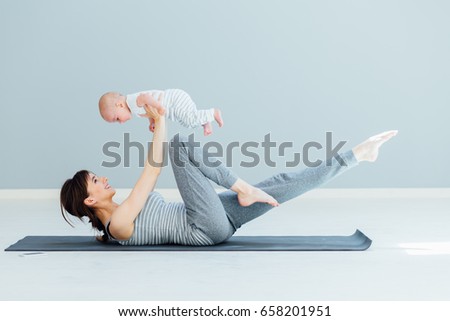 Sport, motherhood and active lifestyle concept - side view of young mother workout together with her baby over gray wall background. Mother having fun and playing with her little son. Royalty-Free Stock Photo #658201951