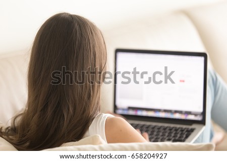 Woman lying on sofa with laptop distantly working from home. Female user browses Internet online, communicates in social network, surfing important information. View over the shoulder close up photo