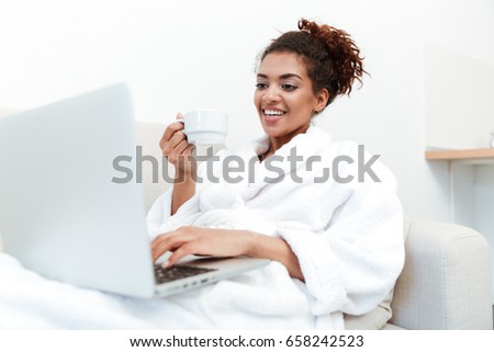 Picture of young african lady sitting on sofa at home and using laptop computer drinking coffee. Looking aside.
