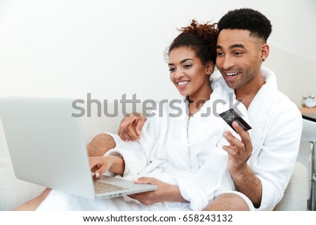 Picture of young smiling african loving couple sitting indoors at home and looking aside. Using laptop and holding credit card.