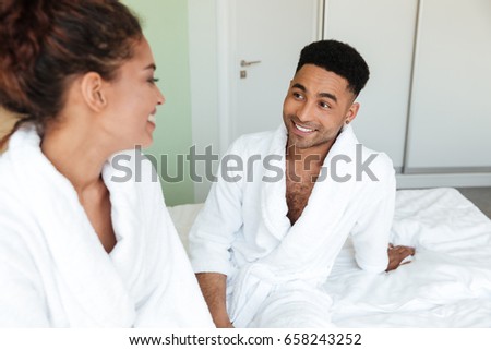 Image of smiling young african loving couple sitting on bed indoors at home and looking aside.