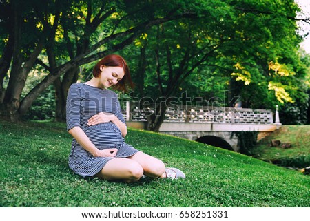 Beautiful pregnant woman in dress on nature, outdoors with copy space. Expectant mother holds hands on belly on natural background of green grass in park. Pregnancy, expectation, new life concepts.