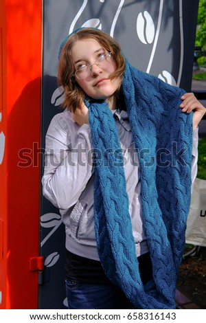 A teenage girl in glasses stands alone in a long knitted scarf