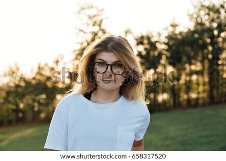 Portrait with analog effect of cute shy american teenager with blonde hair and round nerdy glasses, giggling and smirking into camera, in front of typical house driveway