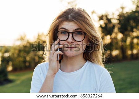 Cute blonde woman with freckles and nerdy goofy funny round glasses chats on phone with her friend or awaits someone to pick up line, in soft yellow summer sunset light