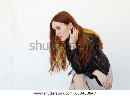Studio portrait of attractive young Caucasian female touching her long red hair and looking away. Headshot of attractive charming young female student having in black leather jacket. Youth and beauty