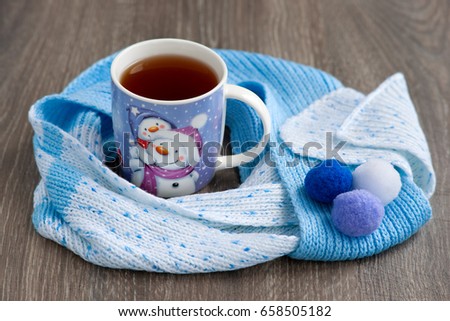 Tea in winter and a scarf. A picture of a snowman on a mug. The snowman's decoration on a tea mug and a knitted scarf create a coziness for the new year. Tea and a warm scarf in the new year warm.