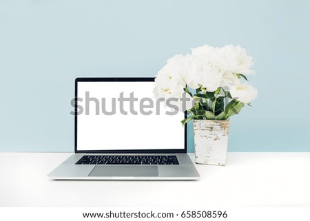 Laptop with white blank screen and flowers in vase on table on blue background. mock up