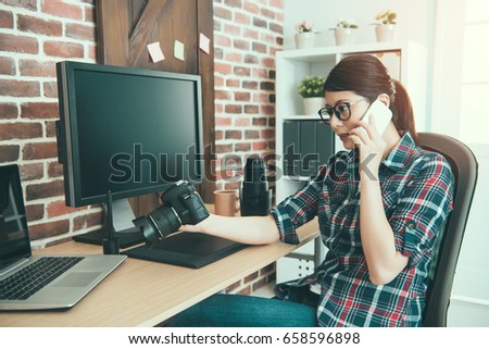 beauty professional female photo company designer holding business camera and calling for photographer checking retouch picture with mobile smart phone sitting on editing desk.