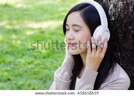 Beautiful asian woman lean on tree listen to music with white headphone in natural park