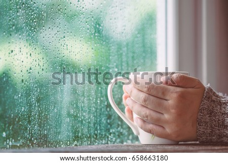 Woman hand holding the cup of coffee or tea on rainy day window background in vintage color tone