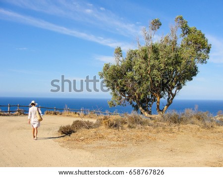Young woman wearing white summer clothes walking on a path leading to the sea. Big tree on the side and blue sea and sky in the background