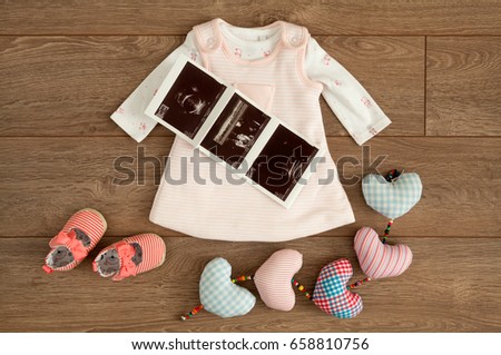 Flat lay of little pink dress and tiny shoes of infant baby girl along with sonography pictures sonograms and colorful cotton hearts on brown wooden surface with copy space. 