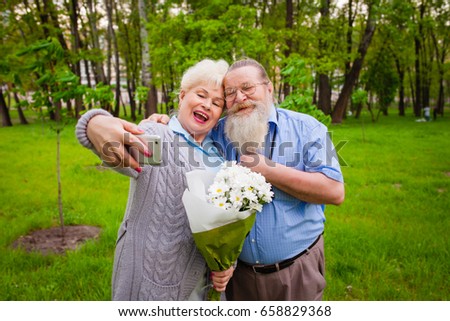 An elderly couple in love stands together in a green park with flowers and making selfie