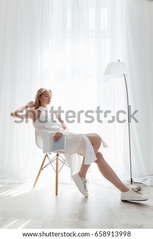 Photo of beautiful blonde lady sitting indoors dressed in white dress. Looking aside.