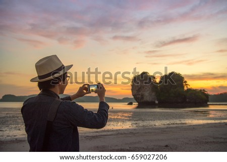 Asian happy tourist man with jean shirt and hat taking photo of beautiful sunset sky on tropical beach island by smartphone. Relaxing on summer holiday. Using mobile phone app on vacation travel.