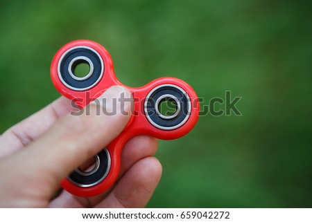 Spinner fidget device in hand.Man holding modern red spinning toy with bearings.Learn cool new balance tricks & have fun with this popular device.Bright green background,focus on gadget