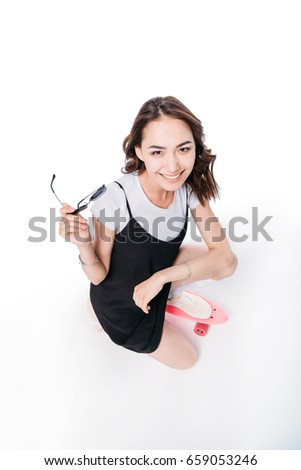 young asian woman holding sunglasses while sitting on skateboard and looking at camera
