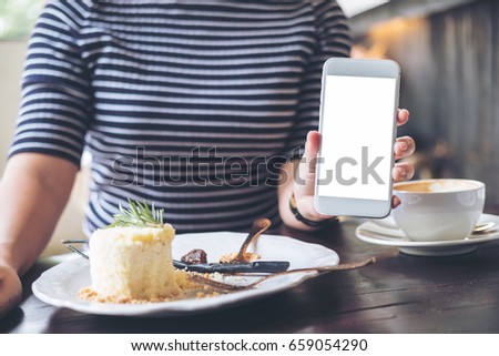 Mockup image of a woman holding and showing white mobile phone with blank screen , cake and coffee cup on wooden table in modern cafe