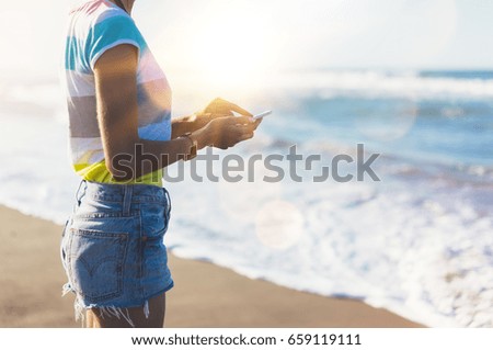 Hipster girl hold on smart phone gadget in sand coastline, mock up of blank screen. Traveler using in female hand mobile on background seascape horizon. Tourist look on blue sun ocean, lifestyle 
