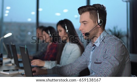 Young handsome men in checked shirt and headset working in call center in modern worldwide office he talking and looking on screen of desktop computer Royalty-Free Stock Photo #659138167