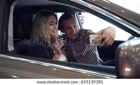 Excited young couple making a decision to purchase a new car sitting inside in a motor showroom checking it out for ownership and make selfie picture using smartphone.
