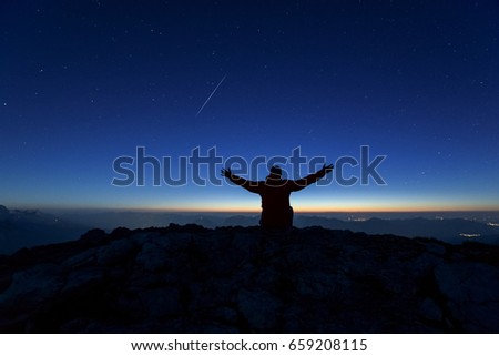 Rear view of a young male hiker at night sitting on a rock near the summit of Westliche Karwendelspitze facing the flat land. Shooting star (meteor) visible in the background. Slight motion blur.
