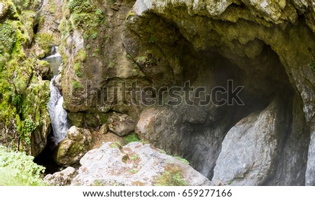 Mountain creek, waterfall and entrance (climb) to the huge Devil's Throat cave in the Rhodope Mountains, abundantly overgrown with deciduous and evergreen forest (Trigradskoto Zhdrelo, Bulgaria) Royalty-Free Stock Photo #659277166