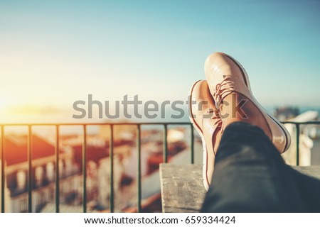 Female feet with glossy pale pink shoes relaxing on wooden table and touristic european city near sea with multiple houses in blurred background with copy space for advertising, logo or your texts