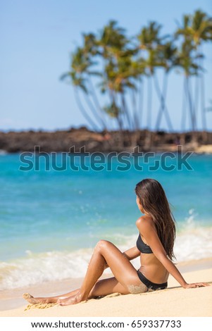 Hawaii tropical beach vacation tourist lady girl sitting down on sand relaxing enjoying sun. Summer travel tourism holiday people on holidays. Bikini woman tanning in summertime.