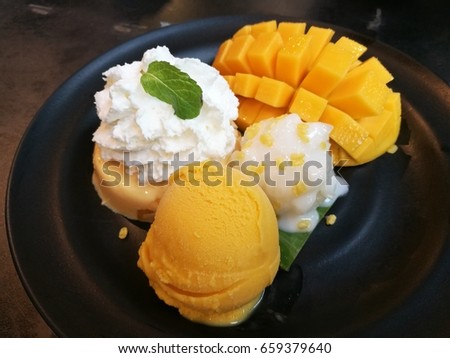 Thai dessert, homemade mango ice cream with fresh sweet mango, whip cream and sticky rice top with creamy coconut milk on black bowl in Thai restaurant, foreground focused with blur background