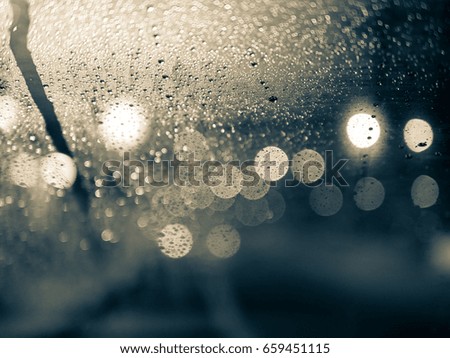 blurred night behind glass window , bokeh lights from traffic street through rain drops on transparent car windshield to the fog city in rainy season