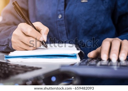 Smart woman hand using pen note information at office with laptop and calculator. Model wearing blue shirt.