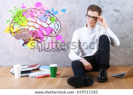 Young businessman sitting on floor with coffee cups, books, laptop and brain sketch on concrete wall. Brainstorm concept