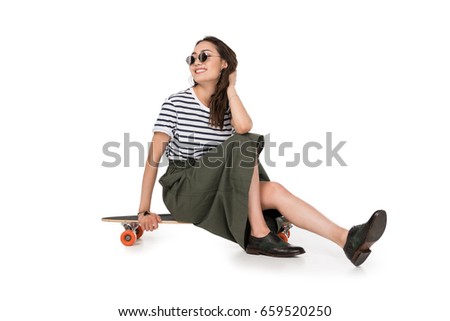 Smiling young woman in eyeglasses sitting on skateboard isolated on white