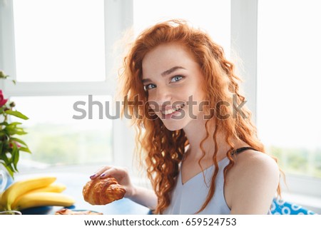 Picture of cute happy young redhead lady sitting at the table indoors near flowers and fruits eating croissant. Looking at camera.