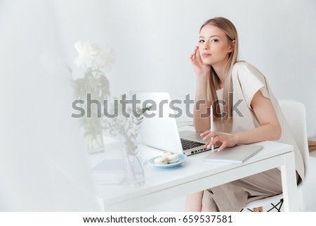 Photo of young serious woman sitting indoors using laptop. Looking at camera.