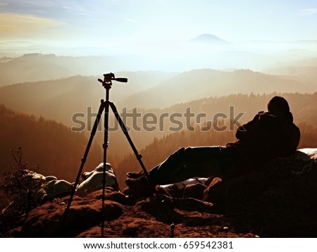Professional photographer takes photos with mirror camera and tripod on snowy peak. Dreamy fogy landscape, spring orange pink misty sunrise in beautiful valley below.