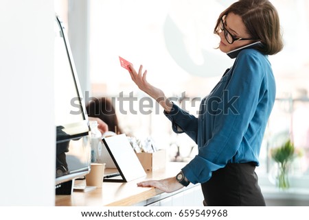 Picture of young woman standing in cafe holding credit card and talking by phone. Looking aside.