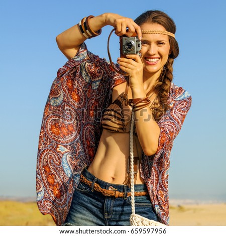 Bohemian vibe vacation. Portrait of smiling stylish gypsy style girl in jeans shorts and cape outdoors in the summer evening taking photo with retro photo camera
