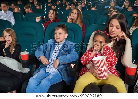Young beautiful woman smiling at her little daughter while watching a movie together at the cinema copyspace people dark auditorium leisure lifestyle enjoyment parenting mother girl pretty.