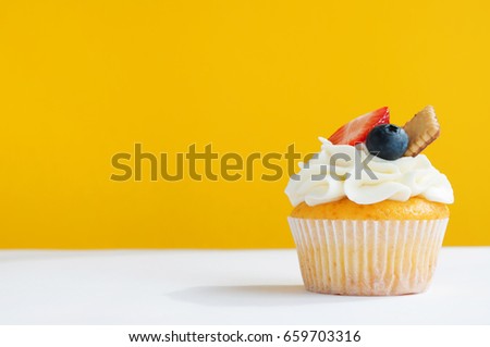 Cupcake with whipped cream, fresh strawberry, blueberry and cookie on yellow white background. The image with copy space. Background for the confectionery menu, cards, greetings, birthday invitations.