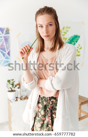 Image of concentrated young caucasian lady painter at workspace. Looking at camera.