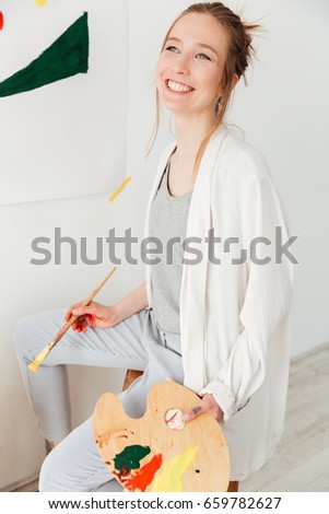 Picture of happy young caucasian lady painter sitting at workspace. Looking aside.